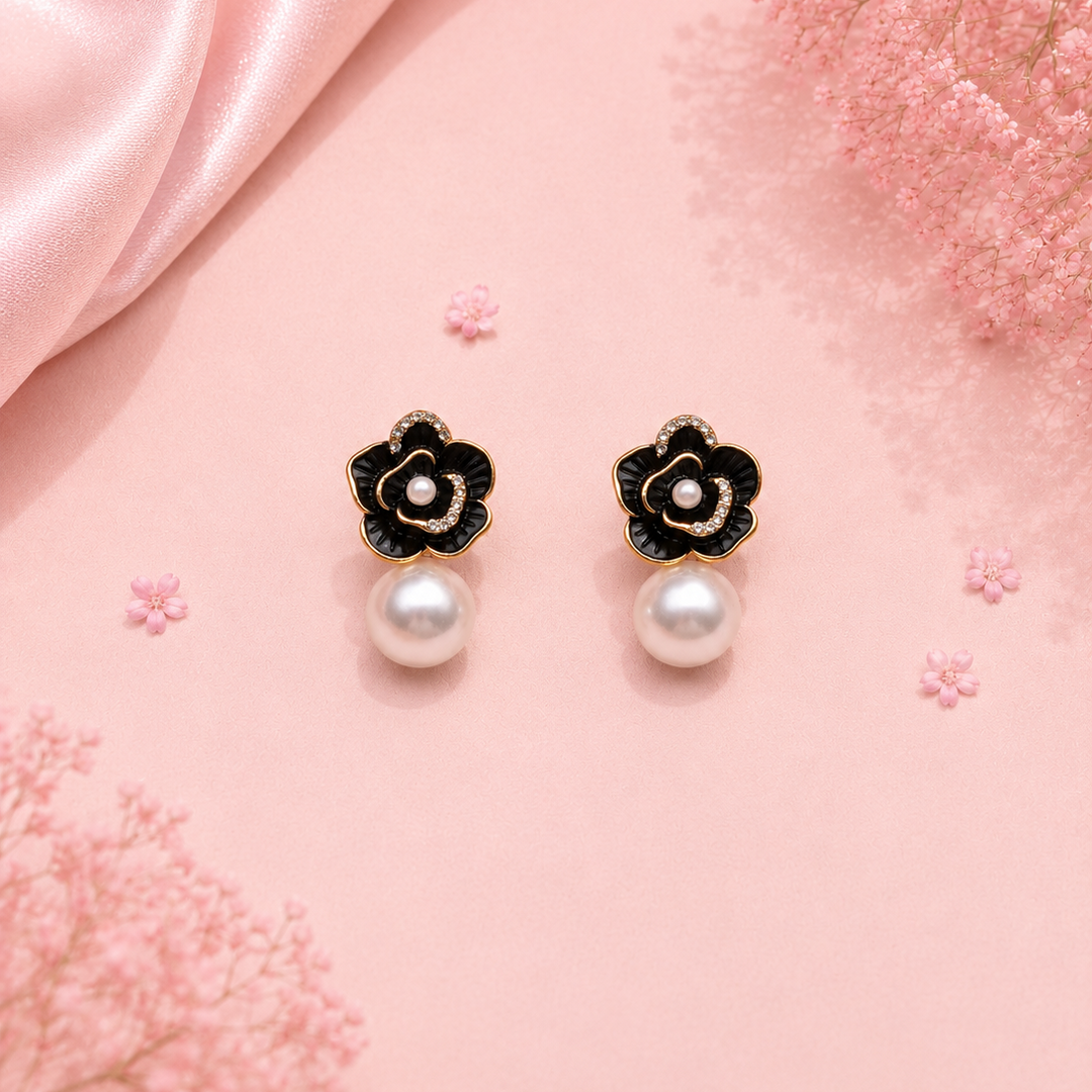 A pair of black and gold flower-shaped drop earrings with white pearl centers and hanging pearls, displayed on a pink satin background with decorative pink flowers.